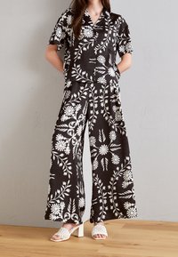 Czarna koszula z krótkim rękawem w kwiatowy wzór oraz pasujące szerokie spodnie. Biały wzór kwiatów i liści. Modelka ubrana w białe sandały na obcasie.
