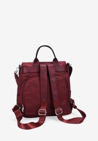 Mochila de tela roja con correas de hombro acolchadas, asa superior y dos bolsillos frontales con cremallera. Presenta una textura suave y costuras detalladas.