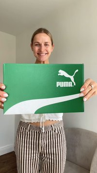 Boîte à chaussures PUMA verte avec un design à swoosh blanc et un logo, tenue par une personne en haut blanc et pantalon rayé, sur un fond neutre.