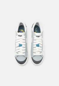 Nike tenisky so sivým koženým zvrškom, bielymi šnúrkami, sivým semišovým detailom a modrým akcentom. Gumová podrážka a polstrovaný golier.