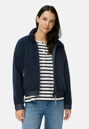 Marineblauwe fleece jas met een rits aan de voorkant, geribbelde manchetten en zoom. Gecombineerd met een gestreept langslavenschirt en denim jeans.
