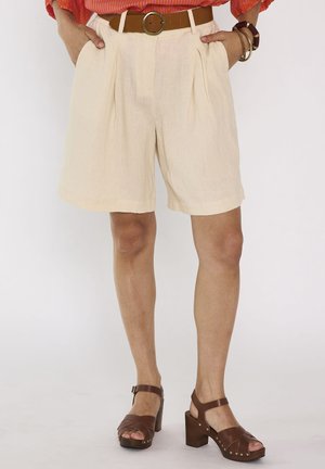 Pantaloncini beige chiaro a vita alta con cintura, indossati con sandali marroni con tacco e top arancione, accessoriati con braccialetti su un polso.
