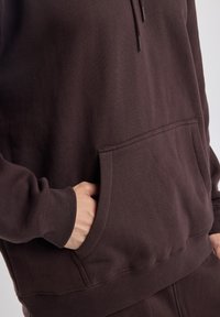 Felpa in cotone marrone con tasca frontale, maniche lunghe, polsini a costine e coulisse regolabile sul cappuccio. Materiale morbido e testurizzato.