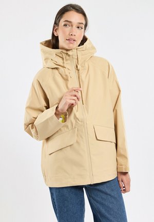 Femme portant une veste beige à capuche avec des poches à rabat à l'avant et un jean bleu, tenant la fermeture éclair de la veste près du visage, regardant sur le côté.