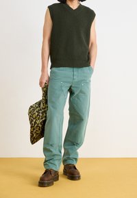 Grön stickad ärmlös topp, teal vidbyxade byxor med synliga sömmar och gulddetaljer, bruna snörstövlar och en leopardmönstrad väska.