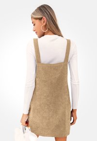 Beige Cordkleid mit breiten Trägern, aus einem strukturierten Stoff gefertigt. Darunter wird ein langärmliges weißes Hemd getragen. Als Accessoires dienen Creolen.