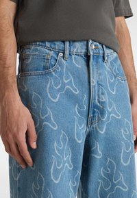 Vaate denim farkut vaaleansinisinä, joissa on valkoiset liekkikuviot, viisi taskua, kupariset yksityiskohdat sekä rento istuvuus vyötäröllä ja jaloissa.