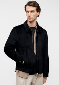 Giacca nera con texture liscia, caratterizzata da collo alto, chiusura con zip e tasche laterali. Indossata sopra una camicia beige con bottoni.