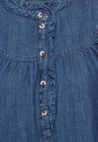 Jeansblus med volang i halsen, knäppning framtill och rynkad söm. Gjord av mjukt blått tyg med en slät textur.