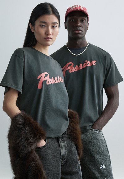 Donkergrijze T-shirts met 'Passie' in rood, gedragen met getextureerde grijze spijkerbroeken; het ene model heeft een bontaccent, het andere draagt een ketting en een pet.