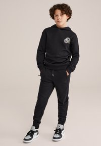 Zwarte sweatshirt met capuchon, voorzien van een cirkelvormige grafiek. Gecombineerd met bijpassende joggers en zwart-witte sneakers met een logo.