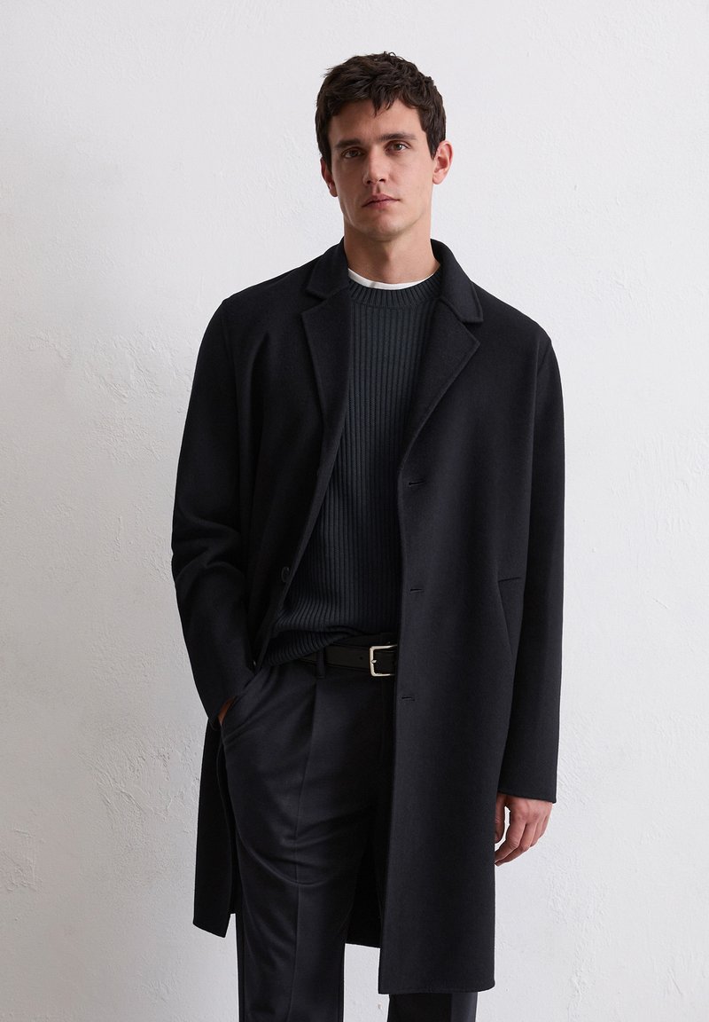 Schwarzer Wollmantel mit klassischem Kragen, gerader Schnitt, Ein-Knopf-Verschluss und Seitentaschen, über einem dunklen gerippten Pullover und dunkler Hose getragen.