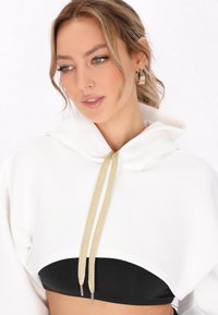 Geknipte witte hoodie met grote capuchon, voorzien van gouden trekkoorden en een zwart hemd zichtbaar eronder. Glad stoffen textuur.