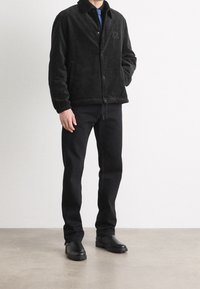 Zwarte corduroyjack met een kraag, drukknopen en elastische manchetten. In combinatie met zwarte broek en robuuste zwarte laarzen. Subtiel merkreclame aanwezig.