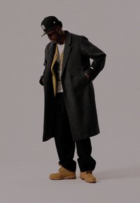 Long manteau en laine gris avec une doublure intérieure jaune, porté sur une chemise blanche et un pantalon noir, associé à des bottes beiges. Silhouette classique et décontractée.