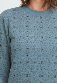 Blauer Pullover mit einem kreisförmigen Muster in Braun, ausgestattet mit einem gerippten Kragen und strukturiertem Stoff. Das Design enthält filigrane Sternmuster.