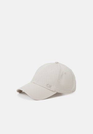 Gorra de béisbol beige con visera curvada, ojales de ventilación cosidos y pequeño logo "CK" en el panel frontal.