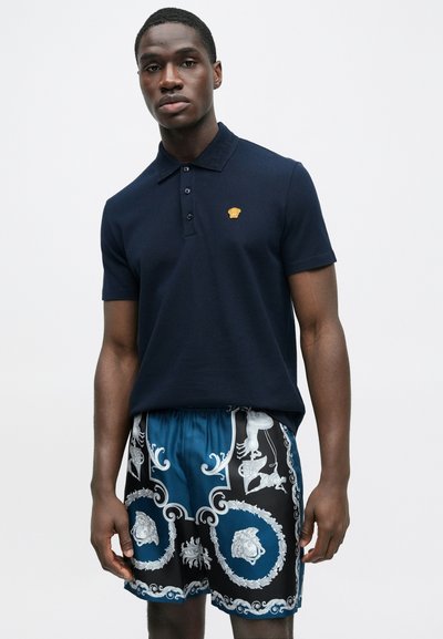 Μπλε σκούρο πόλο μπλουζάκι με χρυσό λογότυπο και μαύρο μαγιό με πολύπλοκο μπλε και λευκό σχέδιο, που προσφέρει άνετη εφαρμογή.