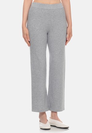 Persona che indossa pantaloni a maglia grigio chiaro a vita alta con gamba larga e scarpe bianche con tacco basso, in piedi su sfondo bianco.