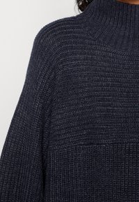 Stickad mörkblå polotröja med ribbat mönster, featuring vida ärmar och en lös passform. Synliga sömmar ger subtila detaljer.
