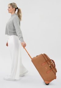 Brauner Rollkoffer mit orangenen Akzenten, Reißverschluss und Rädern, getragen von einer Person in einem grauen Pullover und einer weißen Hose.