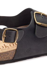 Sandale en cuir noir avec boucles en laiton réglables, semelle en liège et semelle en caoutchouc texturé. Logo embossé sur le côté.