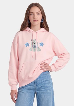 Ung kvinde med langt brunt hår iført blå hoops-øreringe, en pink hættetrøje med en hund og to blå stjerner grafik og lyseblå jeans.