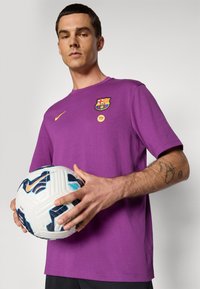 Muškarac u ljubičastoj FC Barcelona majici drži bijelu i plavu Nike nogometnu loptu, stojeći naspram jednostavne svijetle pozadine.