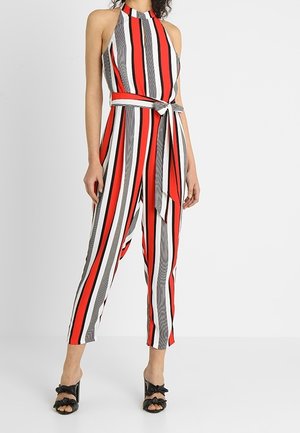 Kvinde iført ærmeløs halter-jumpsuit med lodrette røde, hvide og sorte striber, bindebælte i taljen og sorte åbne tå-hæle.