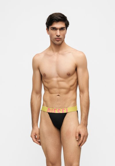 Homme musclé portant un slip noir avec une ceinture jaune portant l'étiquette "Diesel", debout devant un fond blanc uni.