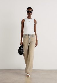 Canottiera bianca attillata, jeans larghi beige chiaro, occhiali da sole e una piccola borsa nera. La modella indossa sandali con tacco bianchi. Sfondo semplice.