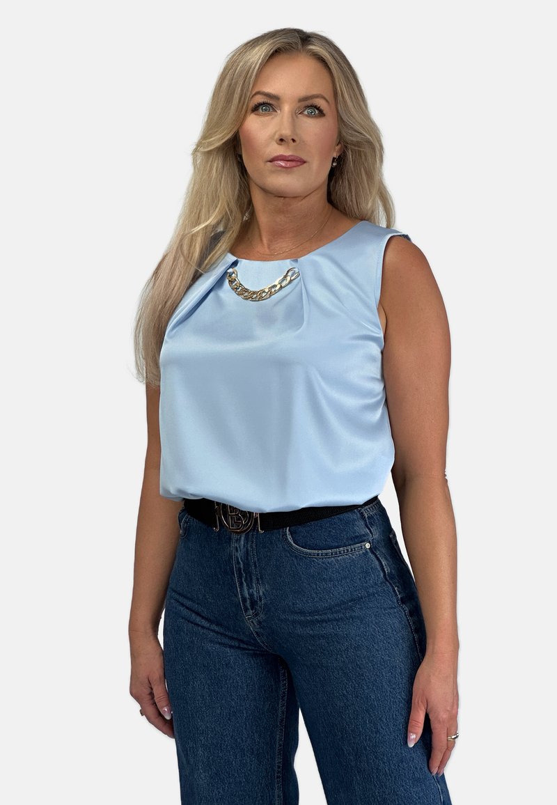 Jasnoniebieski satynowy top bez rękawów z drapowanym przodem i złotym łańcuszkiem na dekolcie, w zestawieniu z ciemnoniebieskimi dżinsami o wysokim stanie.
