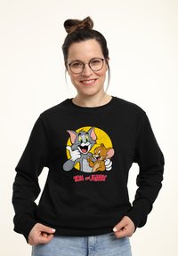 Felpa nera con una grafica colorata dei personaggi dei cartoni animati Tom e Jerry, con uno sfondo a cerchio giallo e testo rosso in basso.