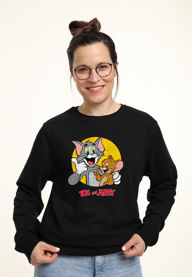Felpa nera con una grafica colorata dei personaggi dei cartoni animati Tom e Jerry, con uno sfondo a cerchio giallo e testo rosso in basso.