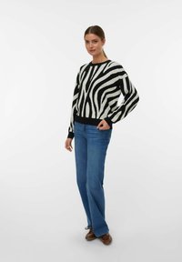 Vrouw in een zwart-witte gestreepte zebrapully, blauwe spijkerbroek en bruine schoenen, staande met één hand in de zak tegen een witte achtergrond.