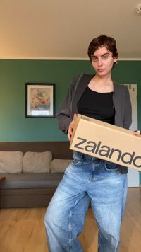 Osobna drži kartonsku kutiju Zalando s velikim crnim tiskanim logotipom. Osoba nosi crni gornji dio i sivi jaknu s patentnim zatvaračem, te izblijedjele plave traperice.
