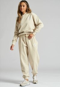Beżowa bluza z kapturem z kieszenią z przodu i logo, w połączeniu z dopasowanymi dresami z elastycznym pasem i ściągaczami na kostkach.