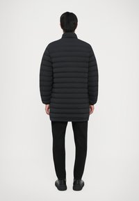 Uomo in piedi con le spalle rivolte, che indossa un lungo giubbotto trapuntato nero, pantaloni neri e sneakers nere, su uno sfondo neutro.