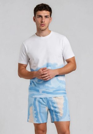 Joven con camiseta blanca con patrón abstracto azul y pantalones cortos a juego con estampado azul y beige, de pie frente a un fondo liso.