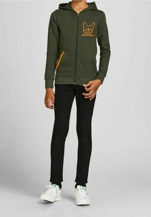 Niño vestido con sudadera verde oliva con cremallera y logotipo de perro, pantalones negros y zapatillas blancas, de pie contra un fondo liso.