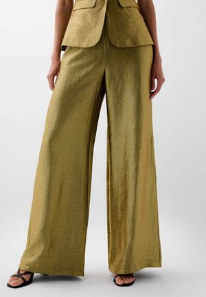 Un pantalon large en tissu métallique olive, avec une finition texturée. La taille est plate, accentuée par des sandales noires à lanières.
