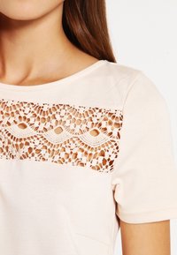 Světle růžové tričko s krajkovým panelem s jemnými vzory, kulatým výstřihem a krátkými rukávy, které vykazuje měkkou, hladkou strukturu.