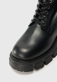 Zwarte leren enkellaars met een ronde neus, voorzien van metalen oogjes en zwarte veters, dikke rubberen zool met diepe profiel voor grip.