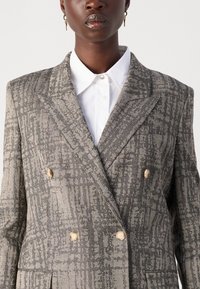 Grau-beige doppelreihiger Blazer mit Hahnentritt-Muster, Notch-Revers und zwei großen hellen Knöpfen. Darunter ein weißes Hemd mit Kragen.
