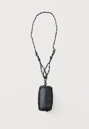 Pochette compacte noire avec fermeture éclair attachée à une sangle de cou réglable noire et blanche à motif sur fond blanc.