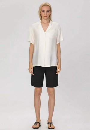 Femme blonde portant une chemise blanche ample à manches courtes boutonnée, un short noir jusqu'aux genoux et des sandales noires, debout devant un fond uni.
