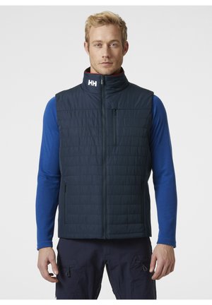 Chaleco aislante azul marino con patrón acolchado, cuello alto, cremallera frontal y bolsillo en el pecho izquierdo. Llevado sobre una camisa de manga larga azul.