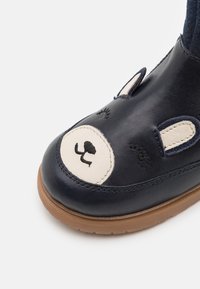 Granatowe skórzane buty z kremowym wzorem w kształcie twarzy misia. Posiadają haftowane oczy i uszy, a także brązową gumową podeszwę dla lepszej przyczepności.