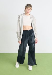 Sudadera gris claro con cremallera sobre una camiseta corta beige, combinada con pantalones negros de corte holgado con el logo de Nike, de pie sobre un suelo verde.