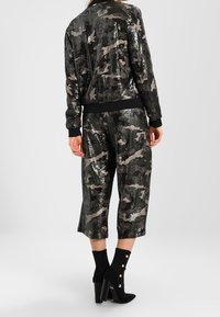 Zwarte en zilveren pailletten bomberjack met camouflagepatroon, gecombineerd met cropped broek van dezelfde stof. Stiletto enkellaarsjes.